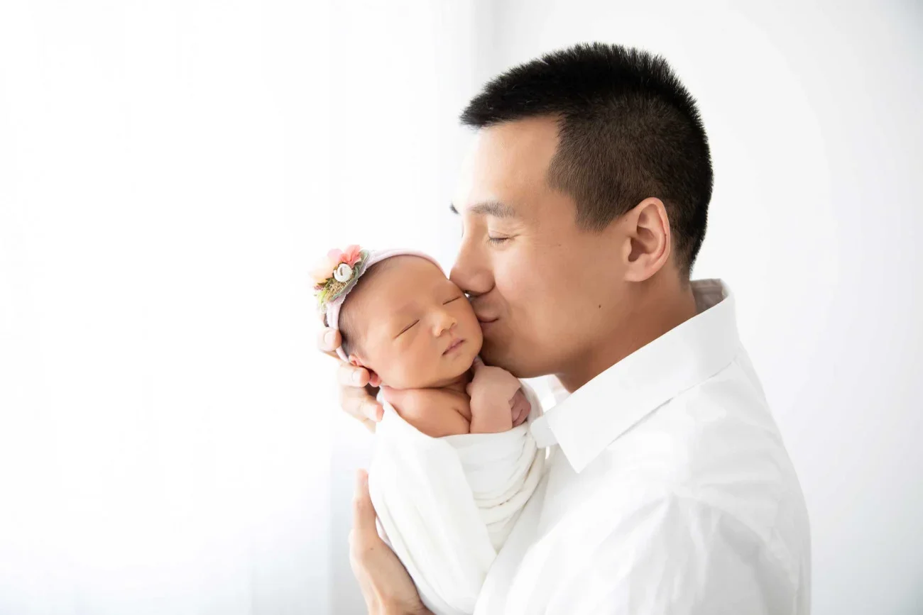 A man holding a newborn baby girl close to his face with eyes closed, touching noses in a gentle kiss, both dressed in white, against a plain white background.