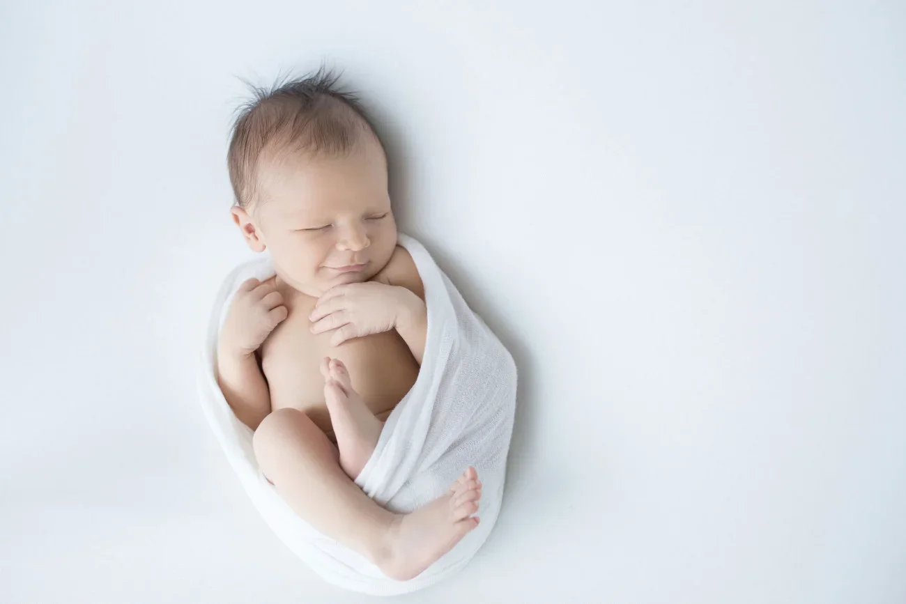 A newborn baby wrapped in a white cloth, sleeping on a white background with a peaceful expression.