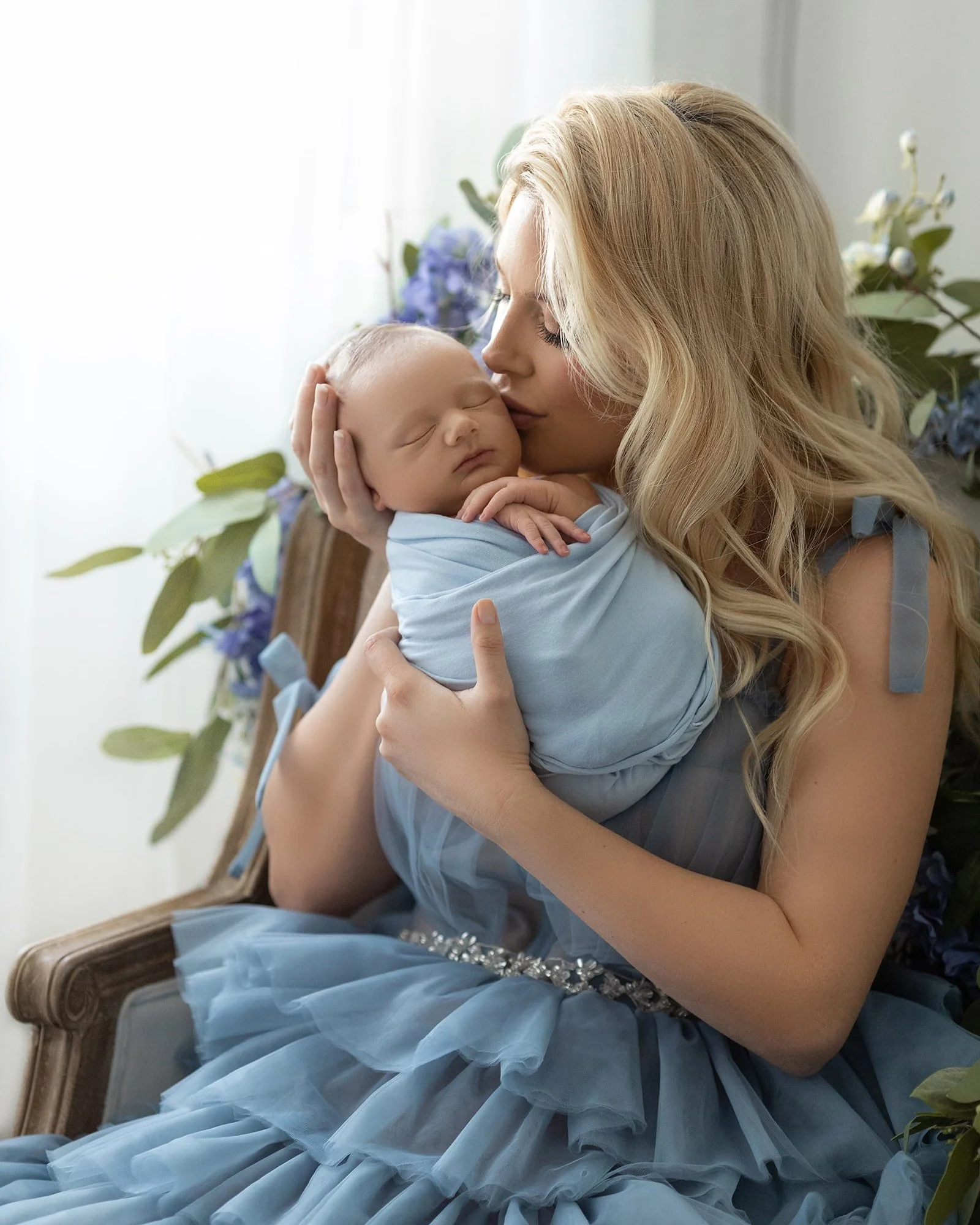A woman with long blonde hair holding a sleeping baby wrapped in a light blue blanket, sitting on a vintage wooden chair with green and purple flowers in the background.