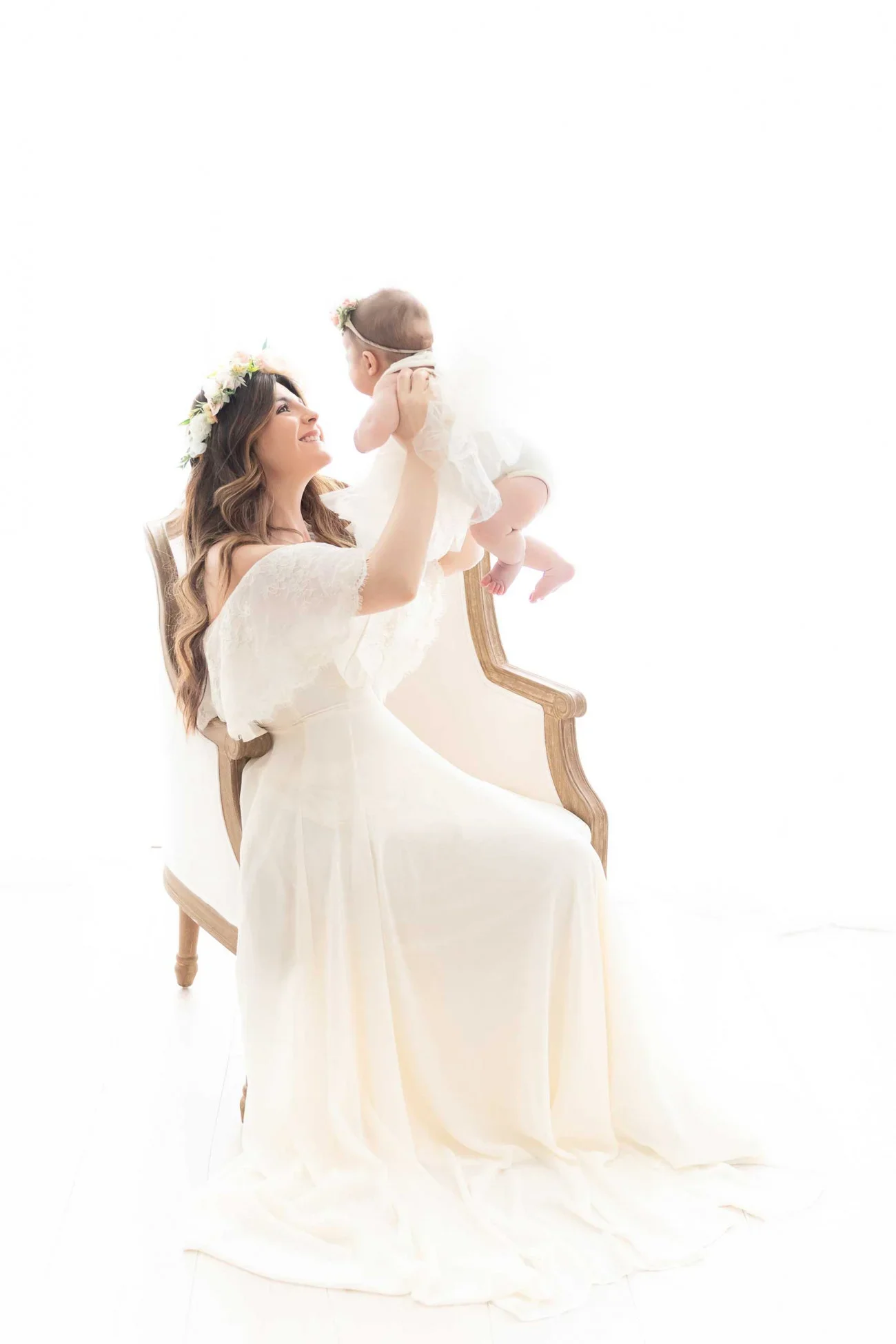 A woman in a white dress and floral crown sitting on a vintage chair, holding a baby girl with a floral headband, against a bright white background.