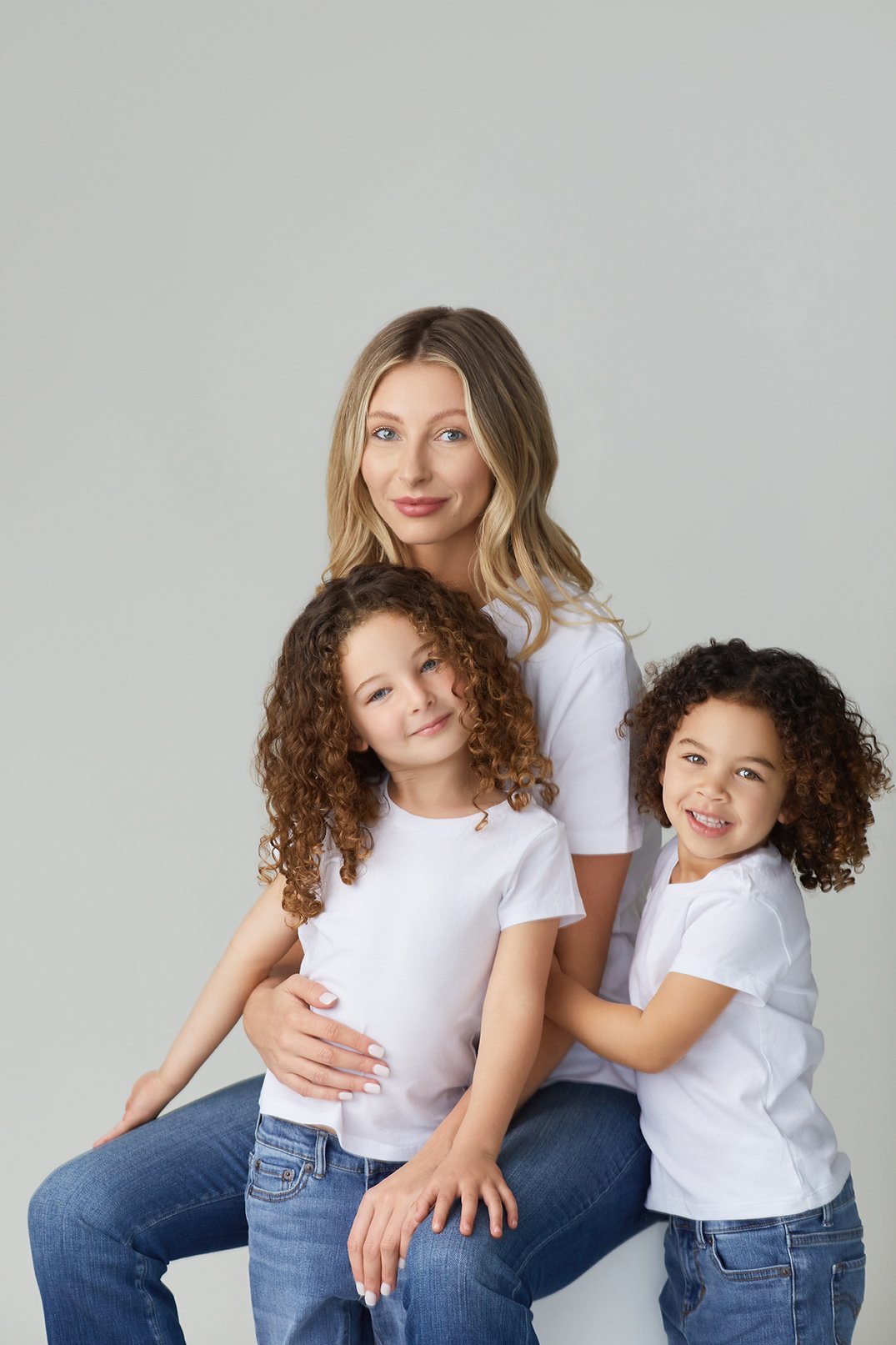 A woman and two young girls, all wearing white T-shirts and jeans, posing together against a plain light gray background.