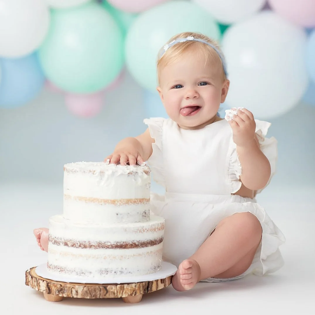 Adorable baby girl with blond hair, wearing a white dress and headband, sitting next to a three-tiered cake with white frosting, holding a piece of cake in one hand and licking her tongue, with pastel balloons in the background.