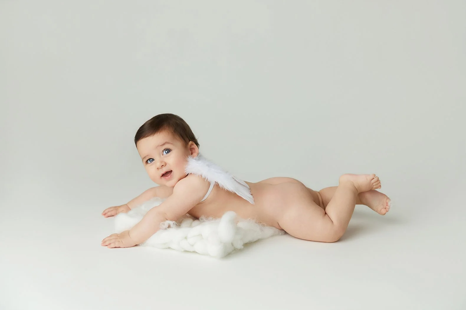 A baby with wings lying on a soft cloud against a plain light background.