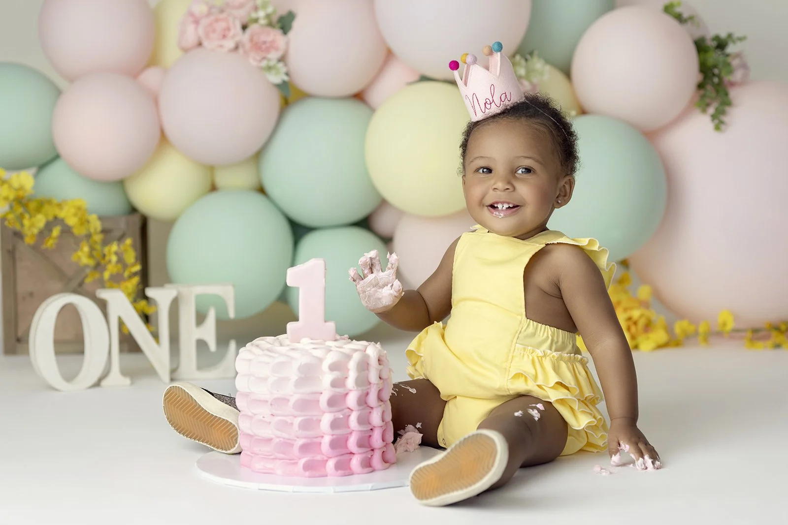 A young girl in a yellow dress sitting on the floor with a pink and white birthday cake in front of her, celebrating her first birthday, with balloons and flowers in the background. She is wearing a birthday crown that says "Nola" and has a big smile