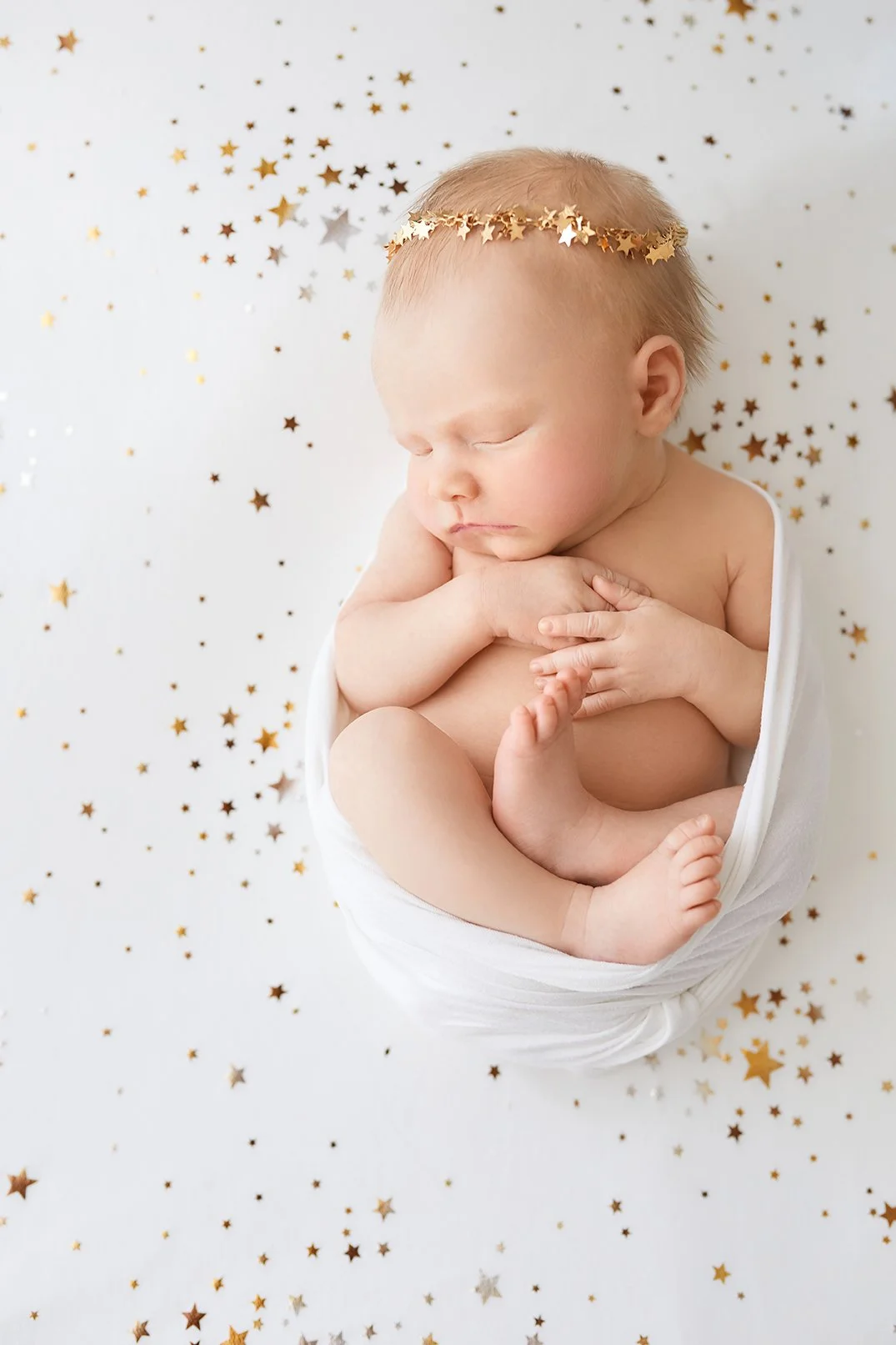 A sleeping baby wrapped in white fabric, wearing a gold star headband, surrounded by gold and silver star-shaped confetti on a white background.