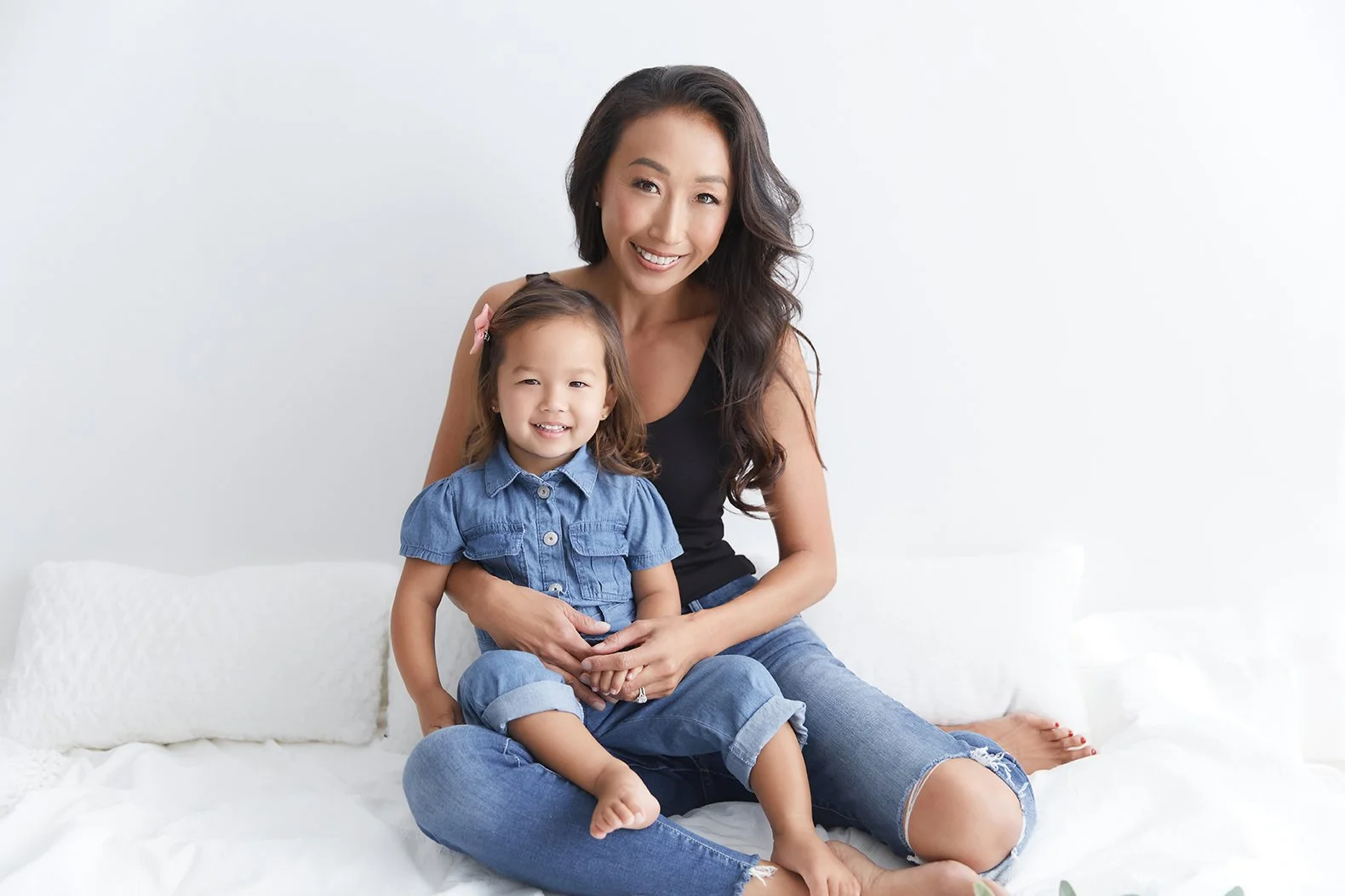 Woman and young girl sitting together on a white bed, smiling at the camera, with a white wall background.