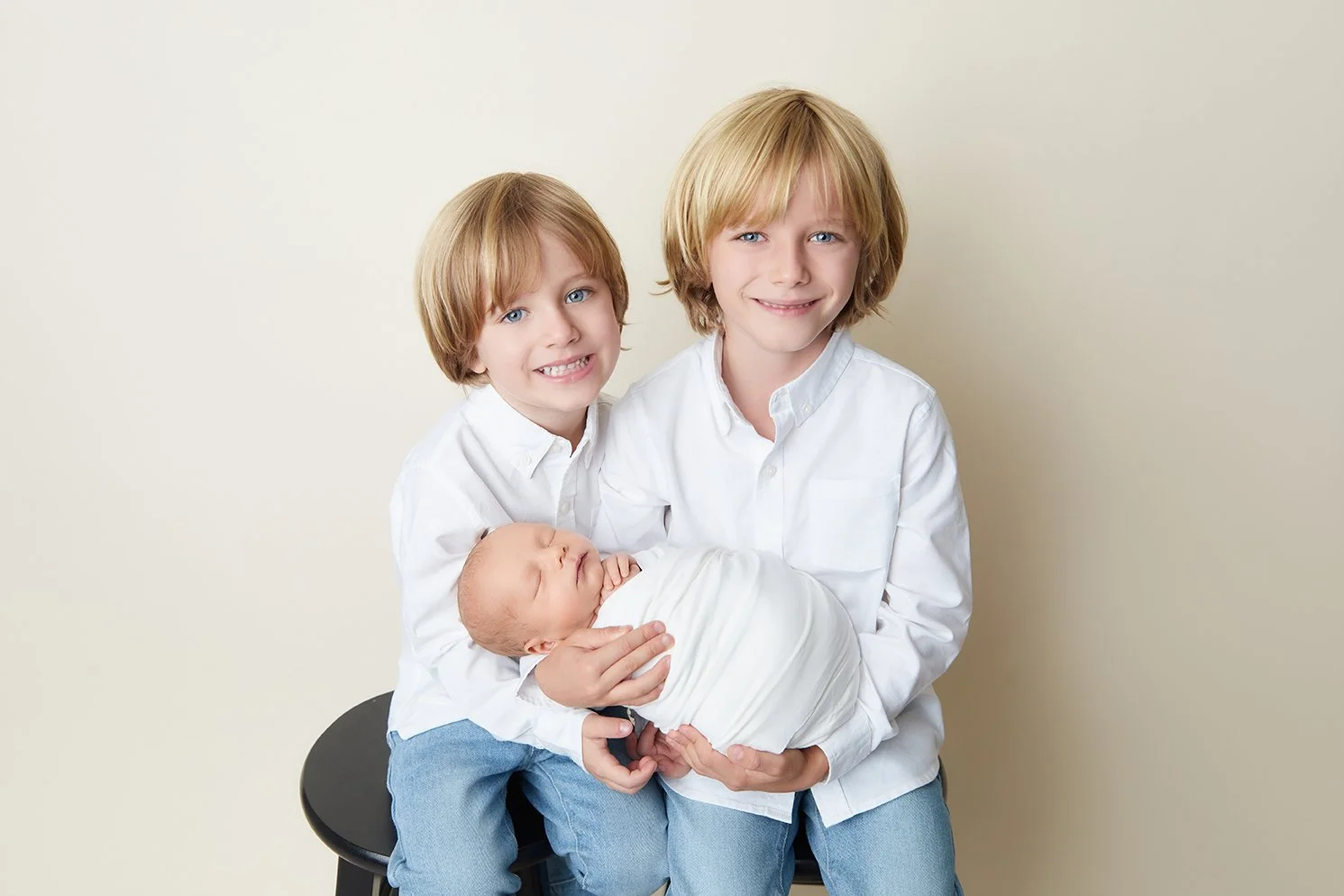 Two young boys with blonde hair and blue eyes, wearing white shirts and blue jeans, smile at the camera while one holds a sleeping newborn wrapped in white cloth, against a plain light-colored background.