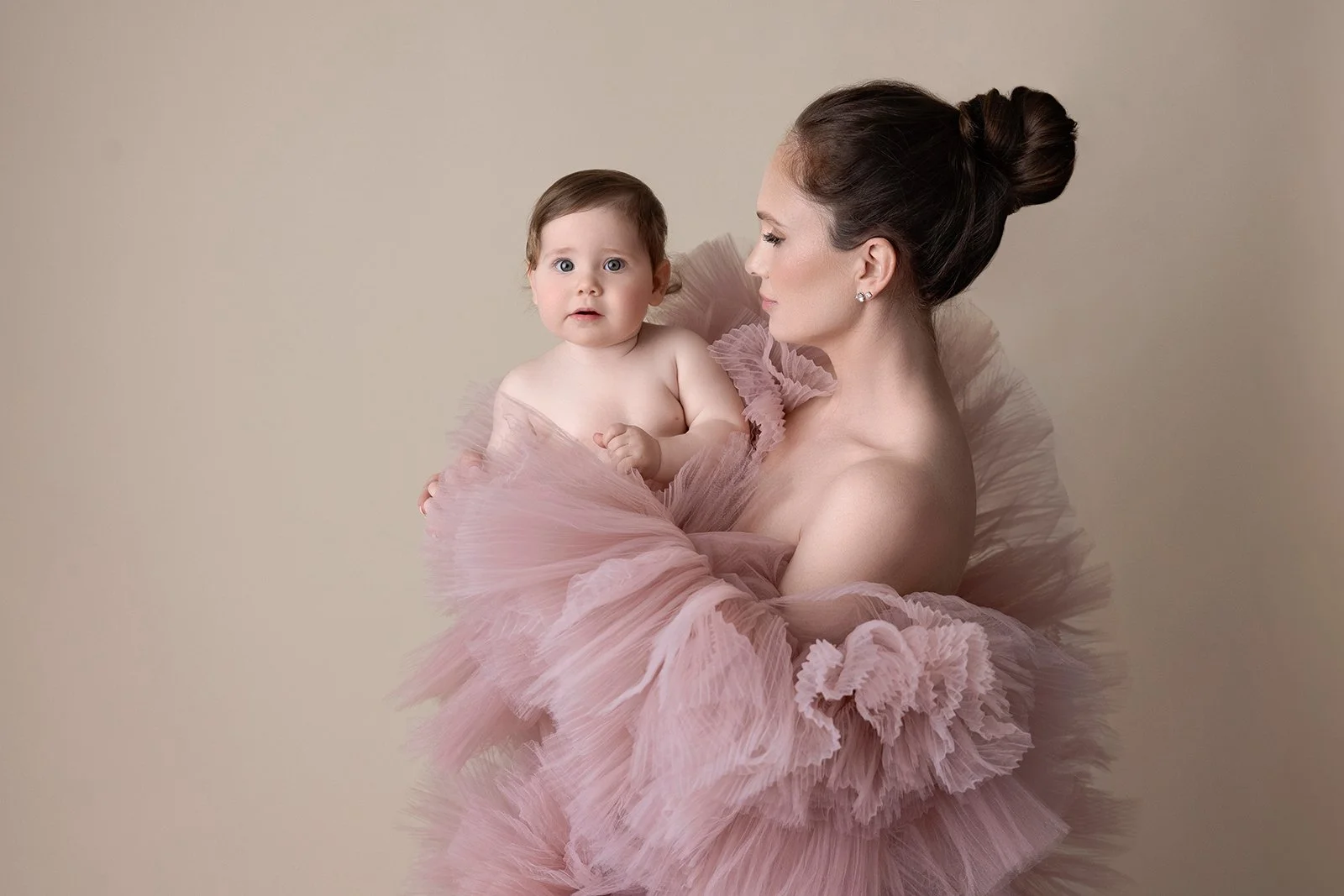 A woman holding a baby girl, both wearing pink tulle dresses, against a plain light background.