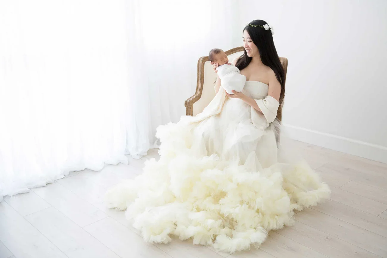 A woman in a white dress sitting on a chair, holding a newborn baby, smiling, in a bright room with white curtains and wooden floor.