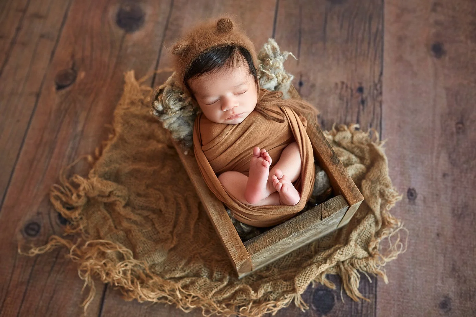 A sleeping baby in a cozy wooden box with brown fabric wrap, wearing a bear hat, on a textured blanket on a wooden floor.