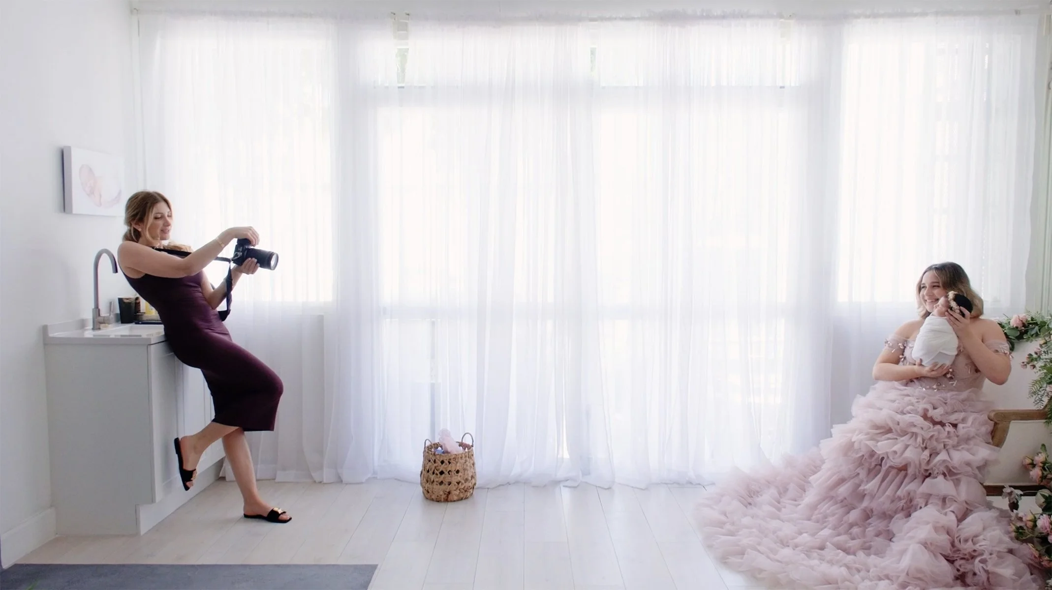 A woman in a light pink ruffled dress holding a dog, smiling while sitting on a chair, and another woman in a purple dress taking a photo with a camera in a bright, white room.