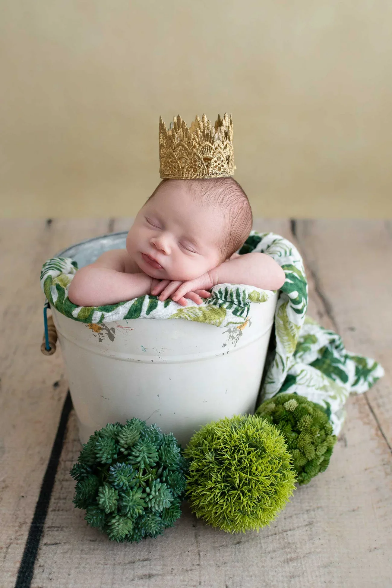 A sleeping baby with a small gold crown on their head, resting their chin on their hands inside a white bucket lined with green and white fabric, surrounded by three green and yellow ball-shaped artificial plants on a wooden surface.