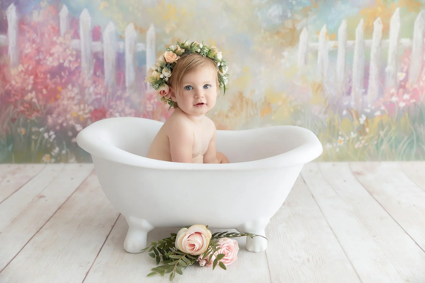 A baby girl with a floral headband sitting in a small white vintage bathtub on a wooden floor with a colorful, nature-themed backdrop and a floral arrangement at the base of the tub.