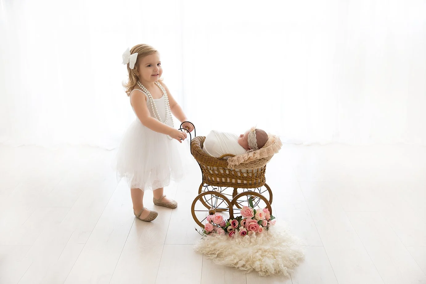 A young girl in a white dress with a large white bow in her hair and string of pearls is pushing a vintage doll pram with a baby doll wrapped in white cloth inside, surrounded by pink roses and cream-colored fur on a white floor with a white curtain 