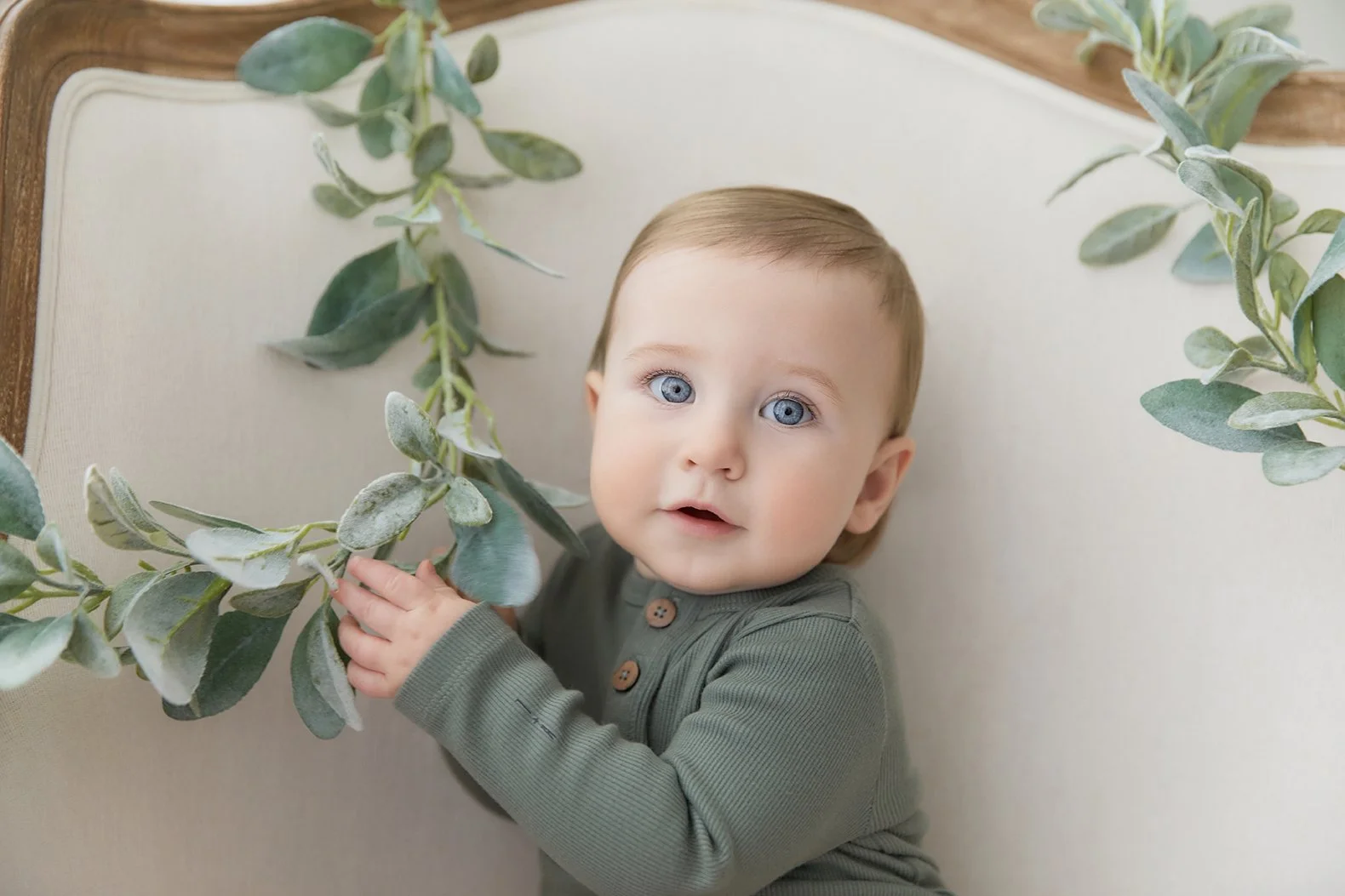 A baby with blue eyes and light brown hair holding a branch of green leaves while lying on a beige cushioned surface.