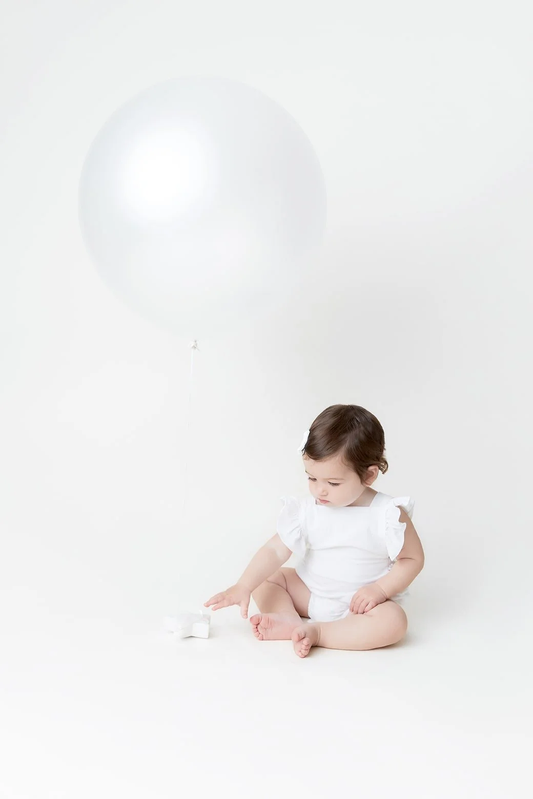 A young girl sitting on the floor in a white dress, playing with a white balloon in a minimalistic white setting.