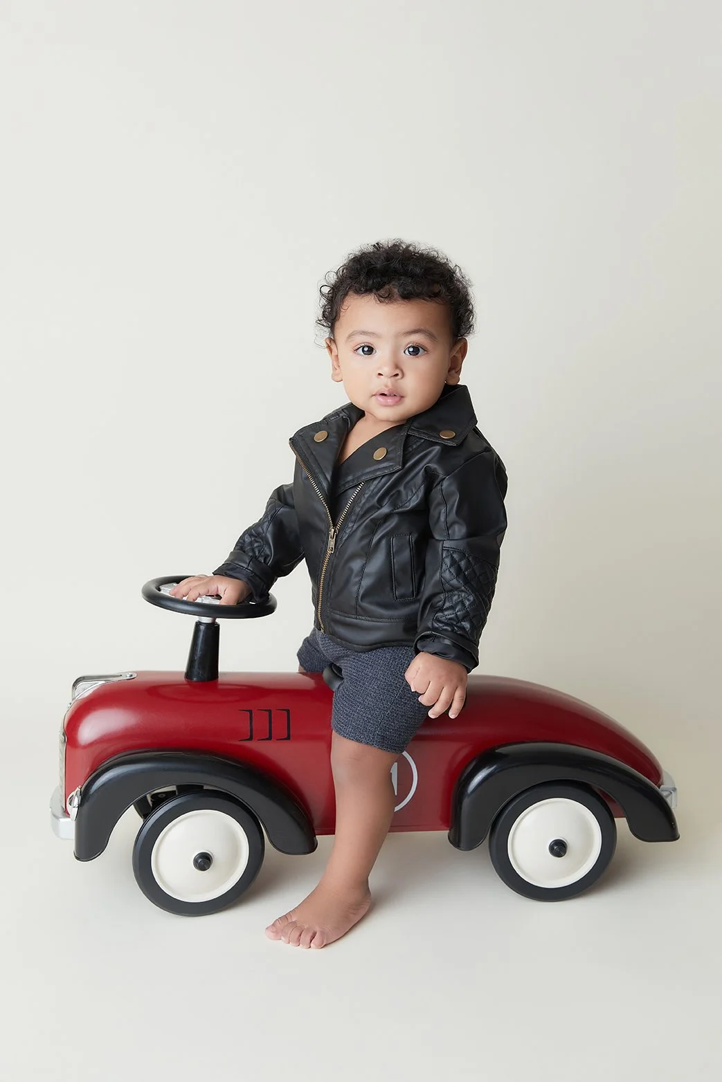 A young boy with curly hair sitting on a red toy car wearing a black leather jacket and gray shorts, looking at the camera.