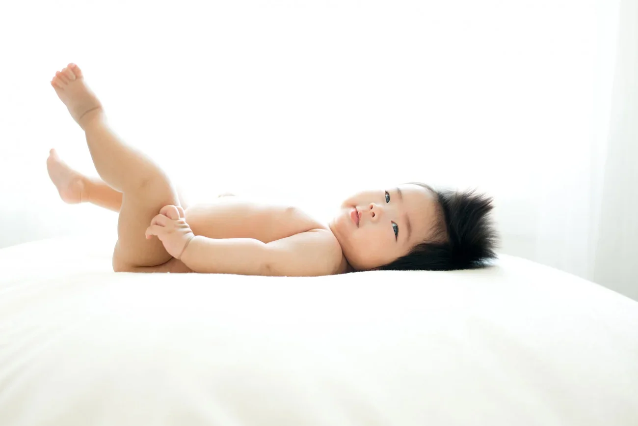 A baby lying on a white bed, looking at the camera, with legs slightly raised, in a bright room with white curtains.