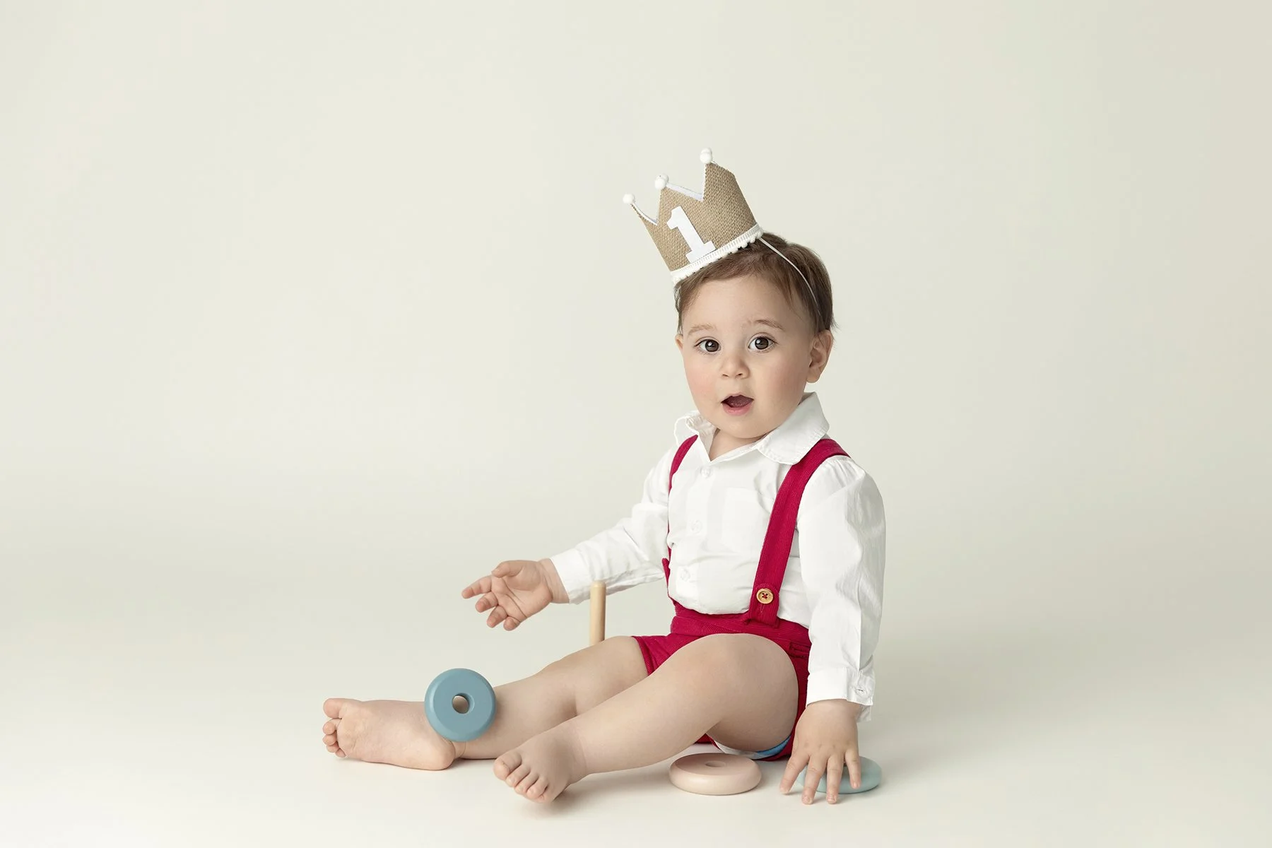 Young child with brown hair, wearing a white shirt and red suspenders, sitting on the floor with large toy rings, wearing a birthday crown, against a plain light background.