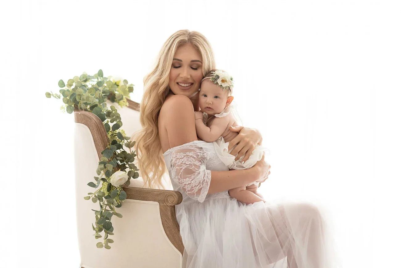 A woman with long blonde hair sitting on a vintage armchair, holding a baby girl with a floral headband, both dressed in white, surrounded by greenery and white flowers.