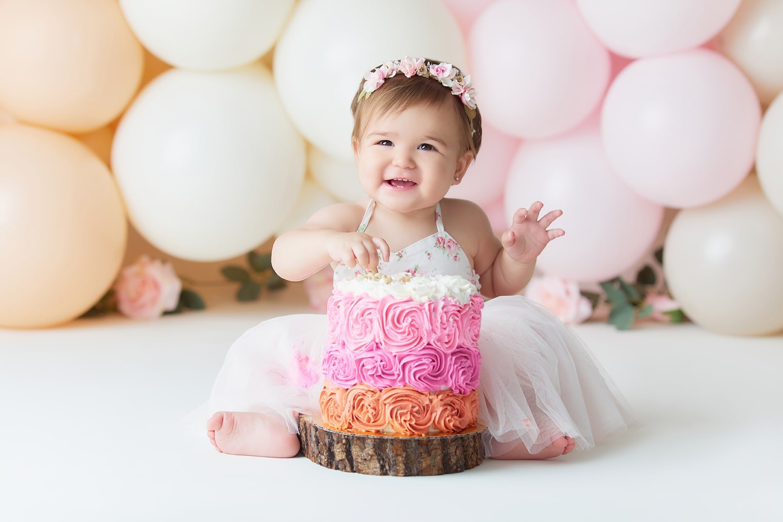 A smiling young girl in a white dress with a pink floral headband, sitting on a white surface with a tiered pink and orange cake in front of her, surrounded by white, cream, and pink balloons in the background.