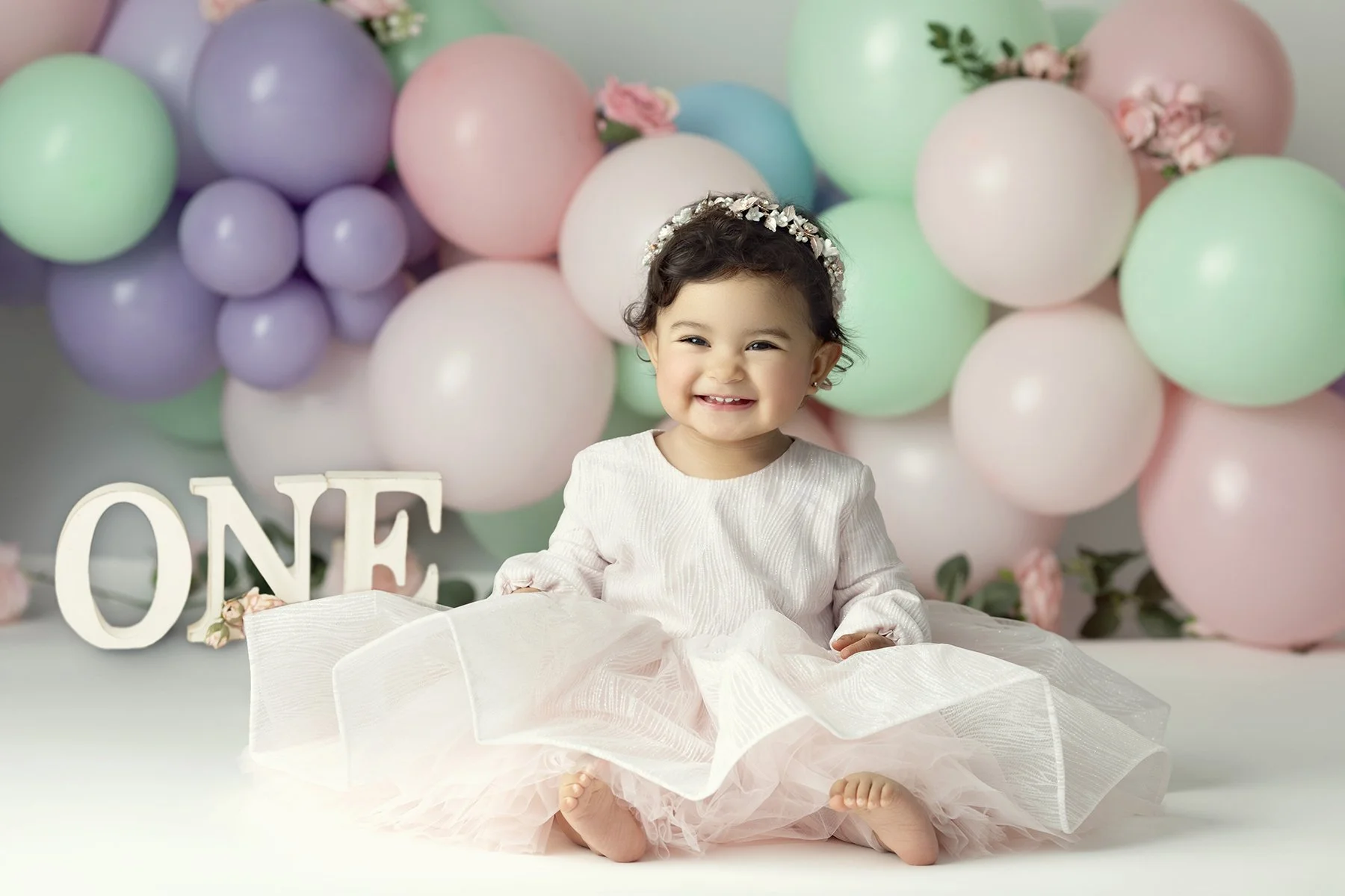 A smiling baby girl with dark curly hair and a floral headband, sitting on the floor in front of pastel-colored balloons and flowers, celebrating her first birthday.