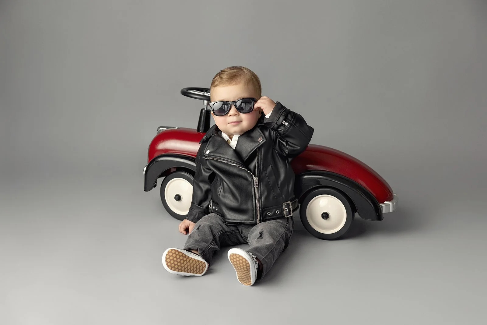 A young toddler wearing sunglasses and a black leather jacket, sitting on the floor next to a red toy car