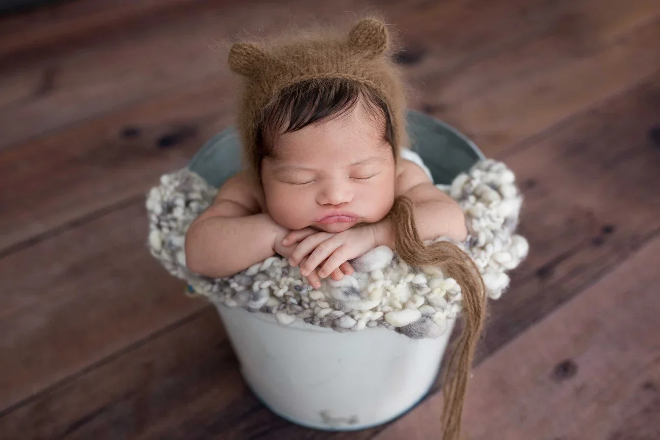 A sleeping baby with a bear hat, resting chin on folded hands, sitting in a white bucket filled with soft, fluffy material, on a wooden floor.