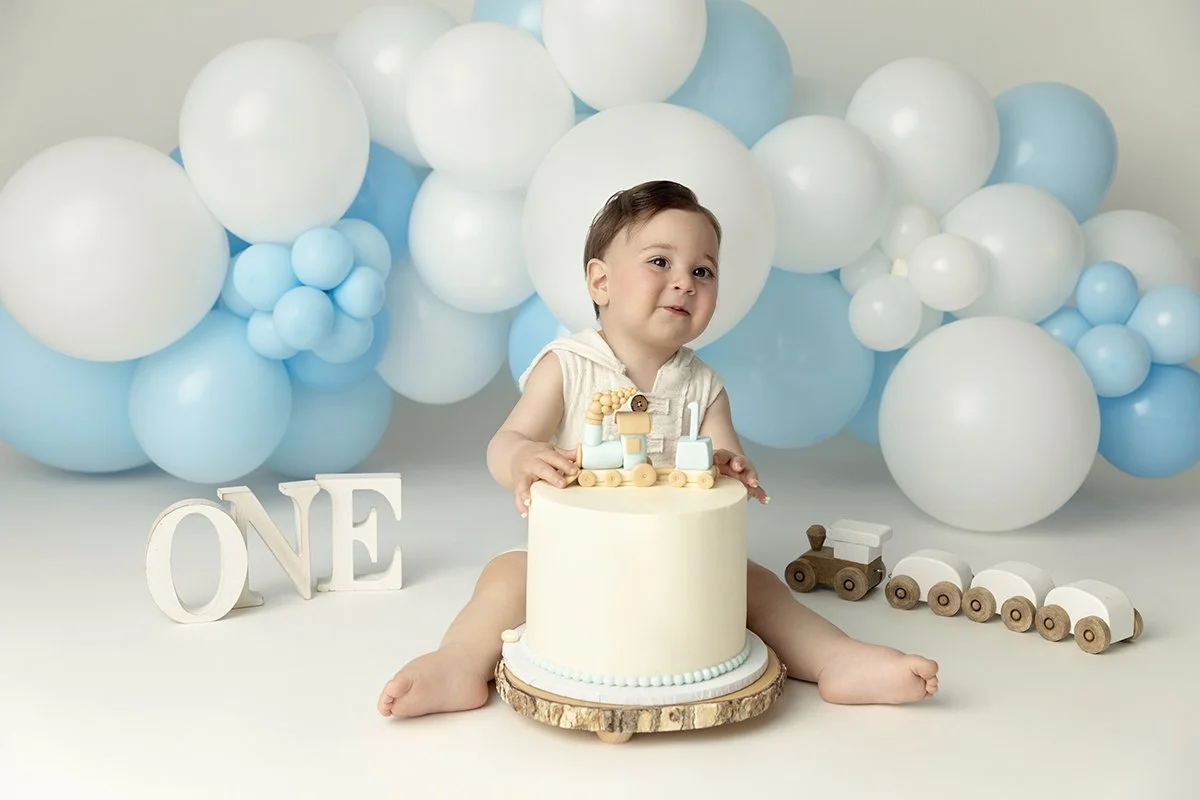 A young child sitting on the floor in front of a white and blue themed first birthday cake with a train topper, surrounded by blue and white balloons and wooden train toys, with a white "ONE" sign nearby.
