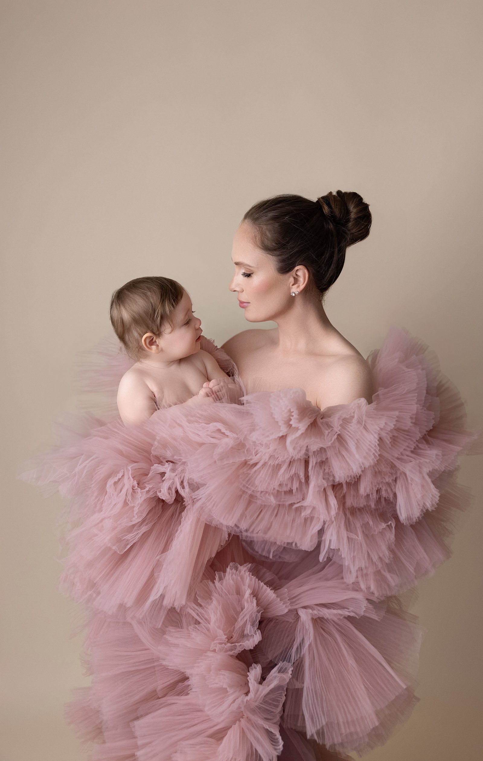 A woman holding a young child wrapped in a pink ruffled dress, both gazing at each other against a plain background.