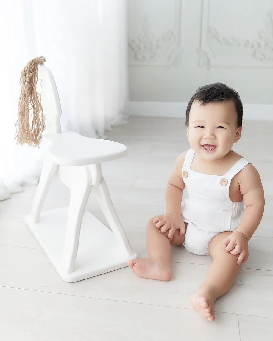 A smiling baby sitting on a light-colored wooden floor next to a white chair with a long, blond wig hanging on its back in a bright, white room.
