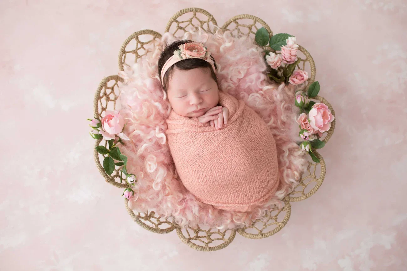 A newborn baby girl sleeping peacefully in a decorative basket, wrapped in a pink blanket, with a matching headband and floral embellishments, on a soft pink background.