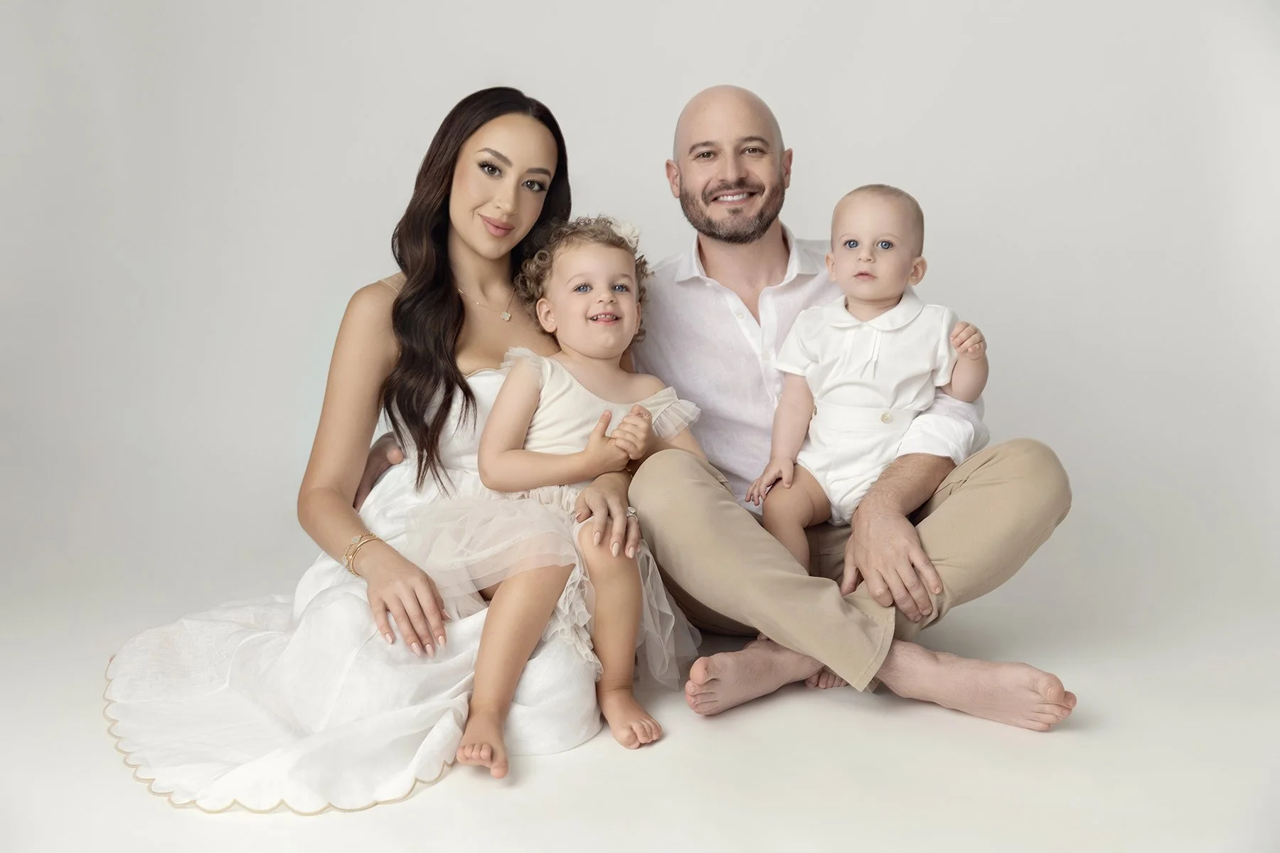 A family of five sitting on the floor against a plain white background. The mother has long dark hair and is wearing a white dress. The father has a beard, is bald, and is wearing a white shirt and beige pants. Their two young daughters are dressed i