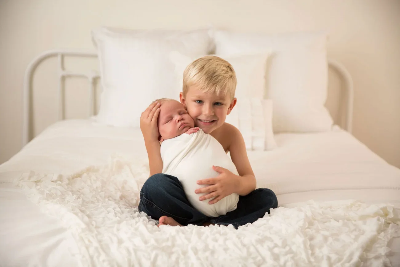 A young boy holding a newborn baby on a bed in a bedroom with white bedding and pillows.