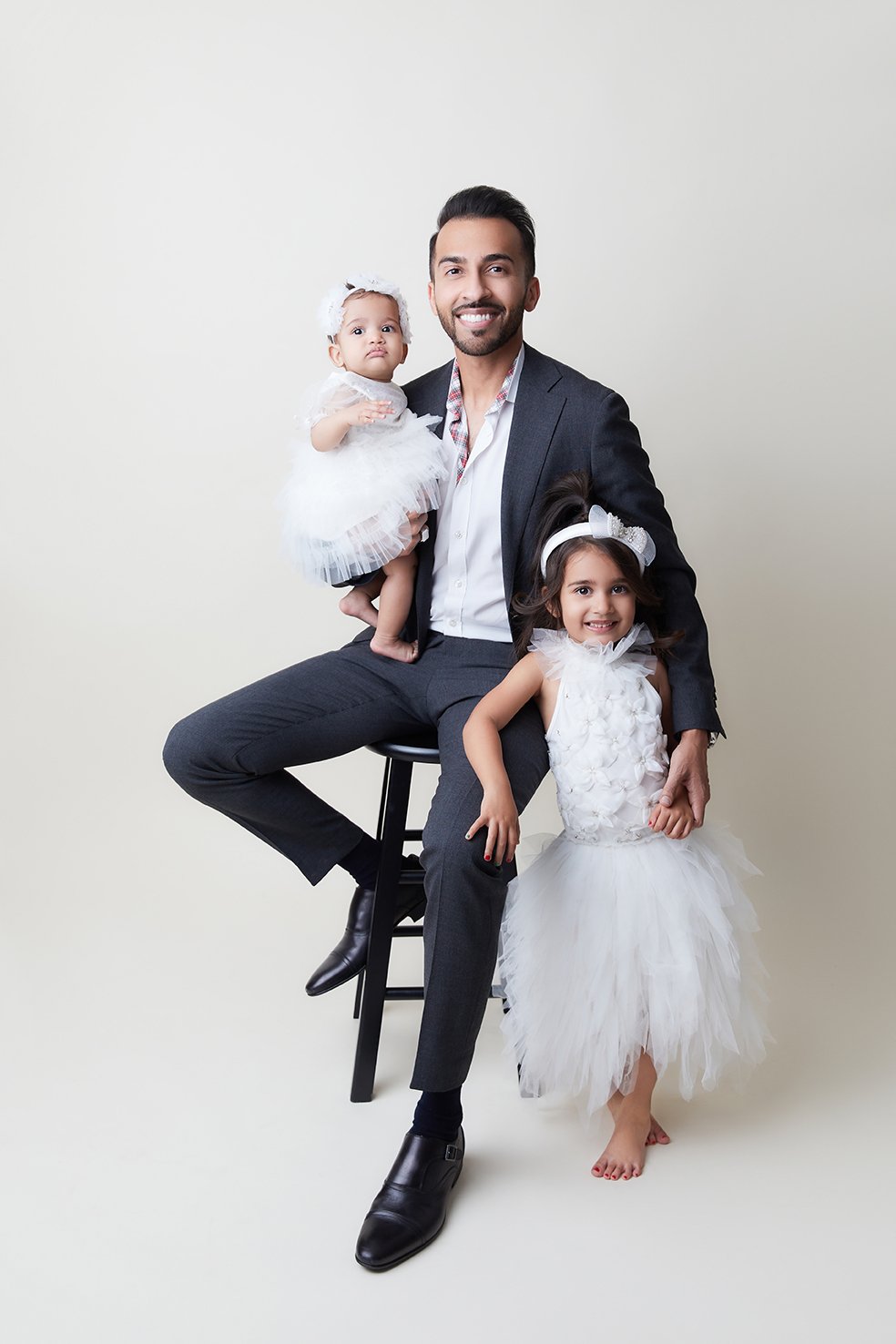 A man in a suit sitting on a stool, holding a baby girl in a white dress, with a young girl in a wedding dress standing beside him, all smiling against a plain background.
