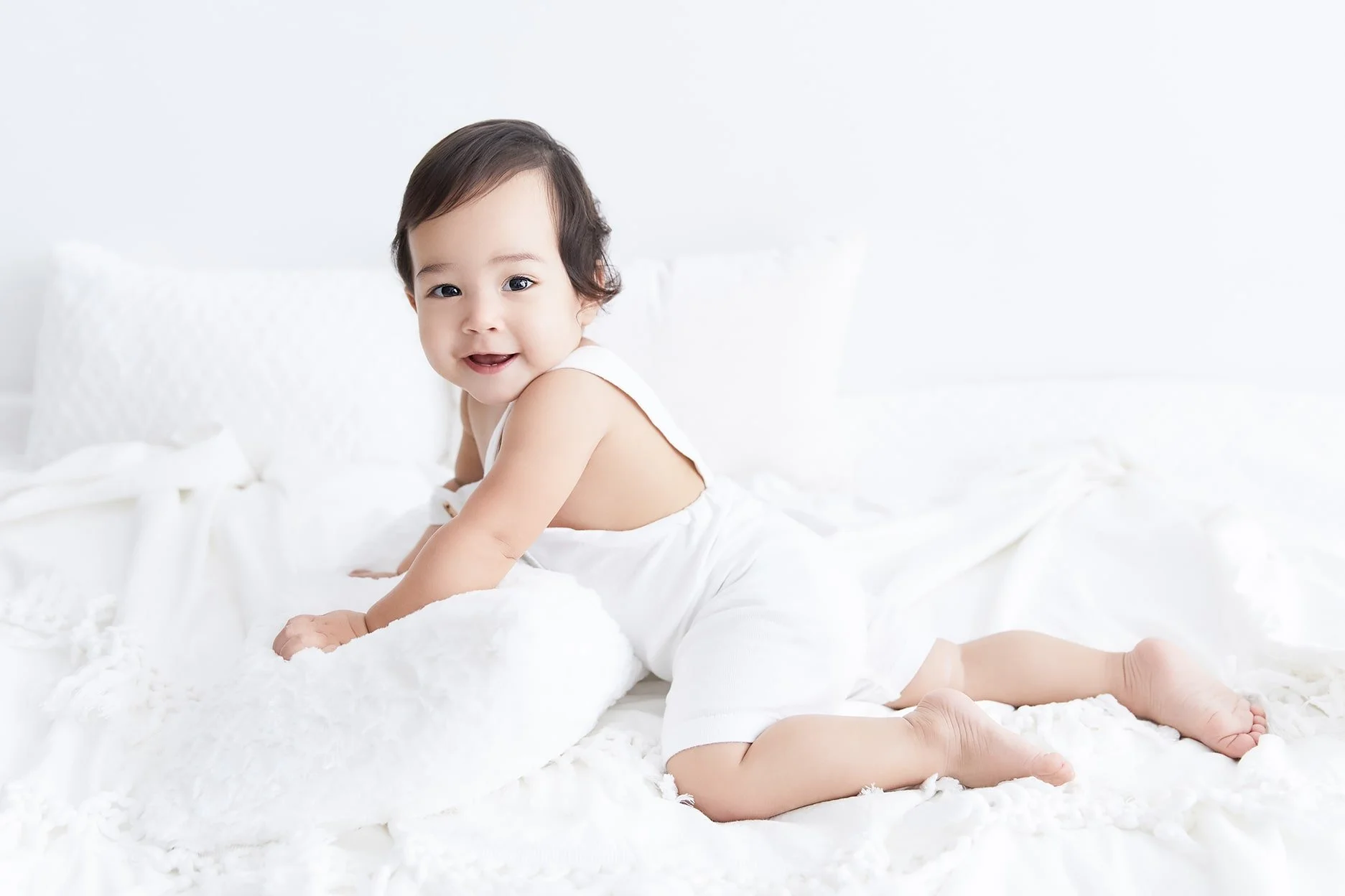 Adorable baby with dark hair smiling while crawling on white bed with white bedding and pillows in a bright room