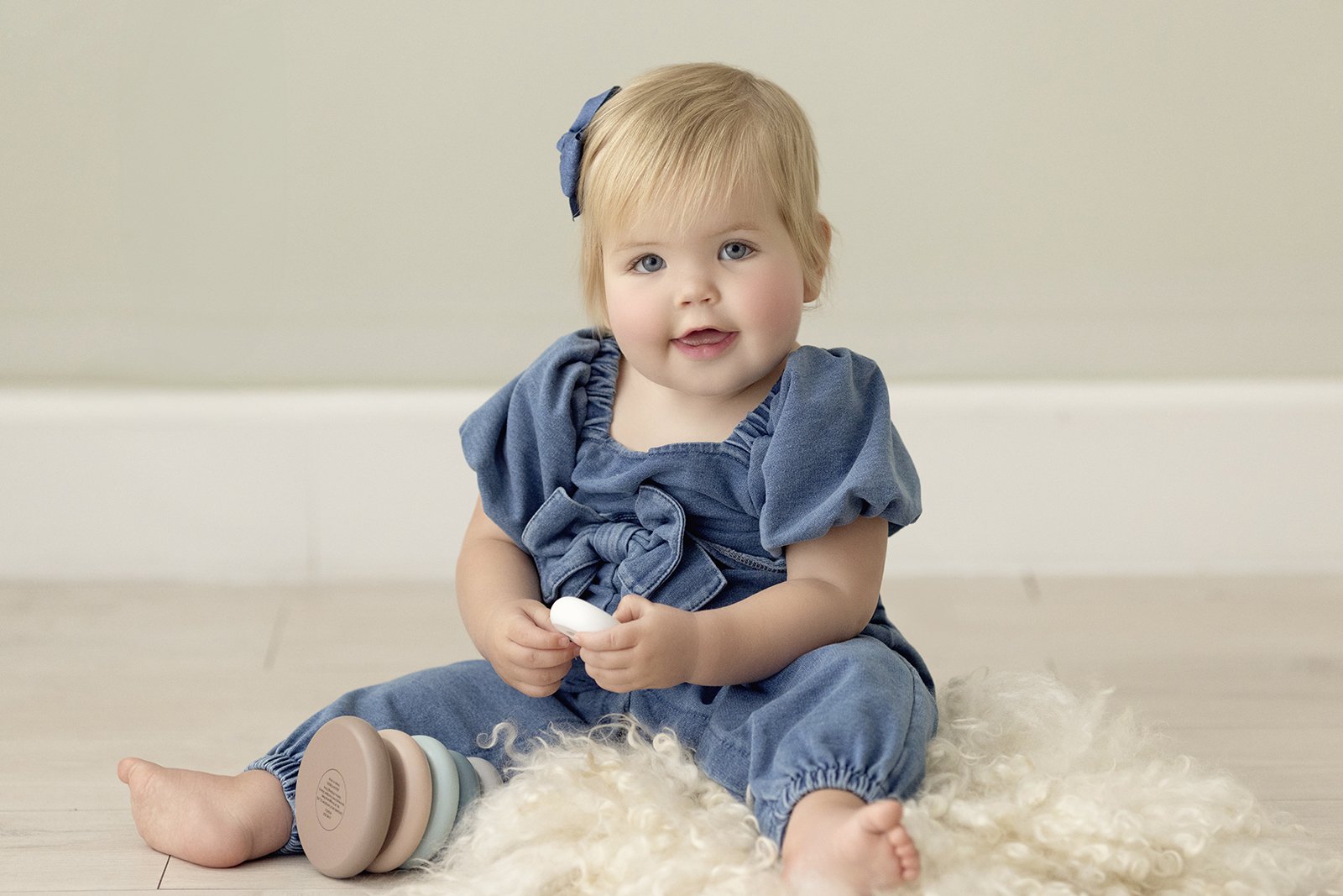 A young blonde girl with blue eyes sitting on a soft white rug, holding a white object, wearing a denim outfit with a matching bow in her hair, and a stacking toy beside her.