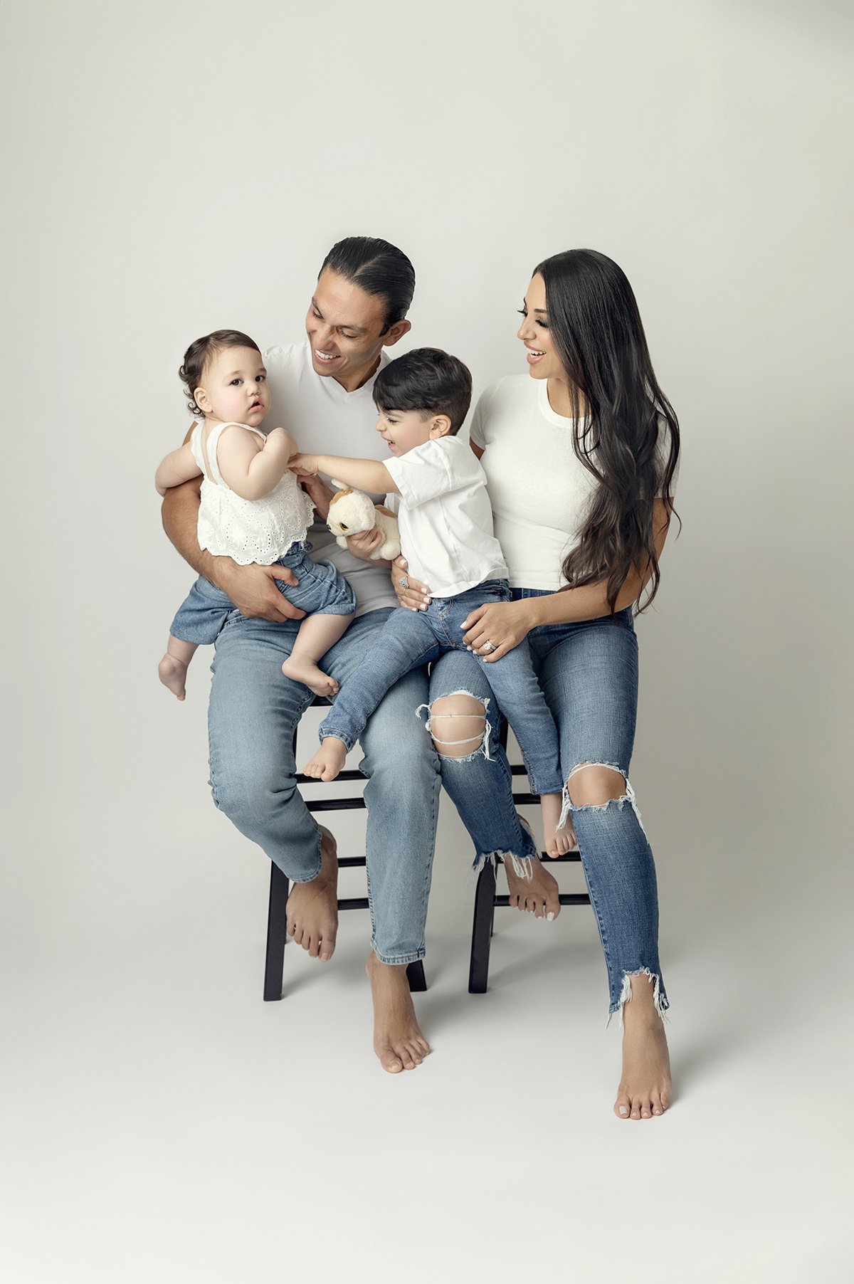 A family of four sitting on a stool against a plain white background, with two parents and two young children playing and smiling.