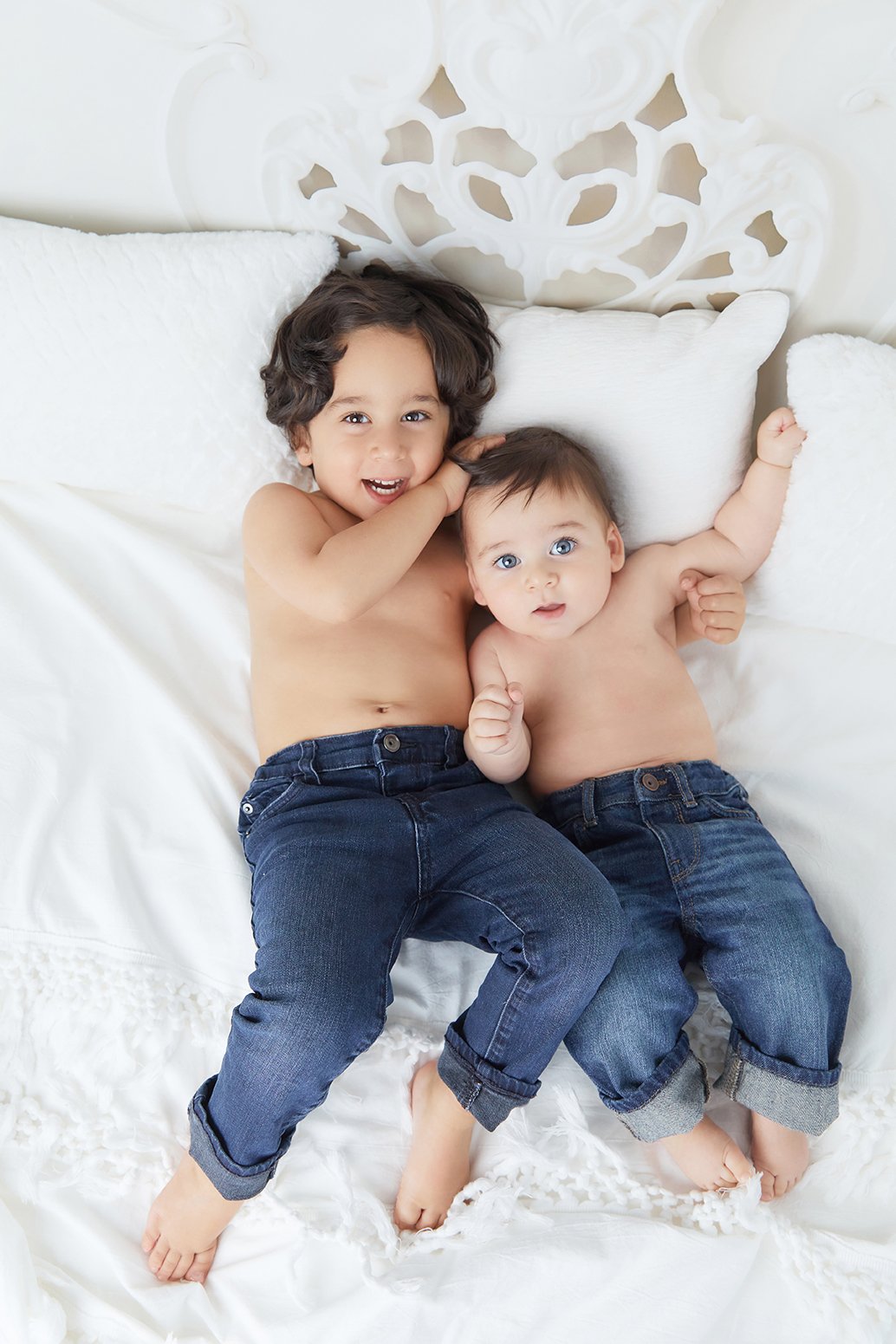 Two young boys lying on a white bed, wearing jeans, smiling and looking at the camera.
