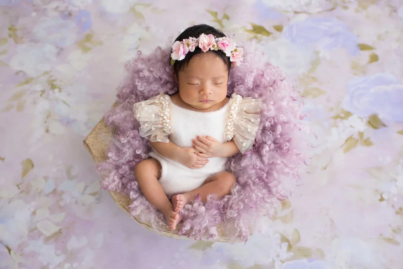 A sleeping baby girl with a floral headband, lying on a soft pink and purple fluffy blanket, surrounded by a floral-themed background.