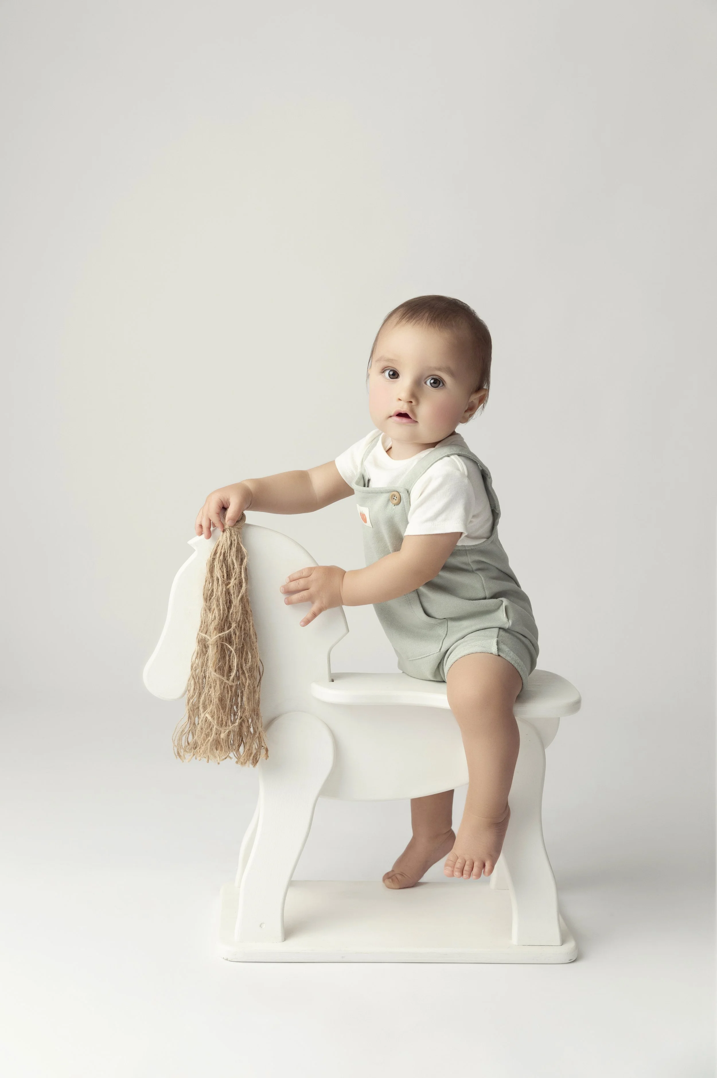 A baby sitting on a white wooden horse-shaped seat, holding a brown mop with a cloth body, against a plain light background.