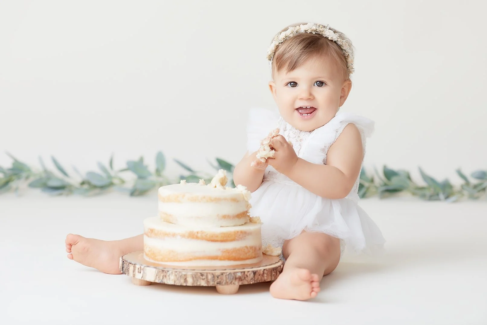 A happy baby girl wearing a white dress and a floral headband, sitting on the floor with a semi-naked cake in front of her, holding cake in her hands, with a white background and greenery in the distance.