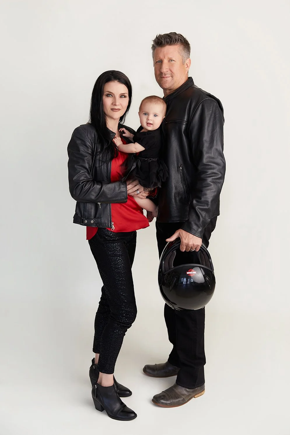 A family of three, a woman, a man, and a baby girl, standing together against a plain background. The woman has long black hair, wearing a black leather jacket, red blouse, and black patterned pants. The man has short gray hair, wearing a black leath