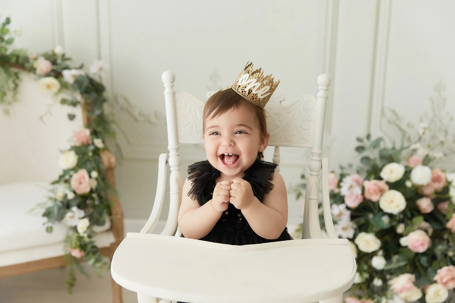 A happy baby girl sitting in a white high chair, wearing a black dress with ruffled sleeves and a gold 'One' birthday crown, with floral decorations in the background.
