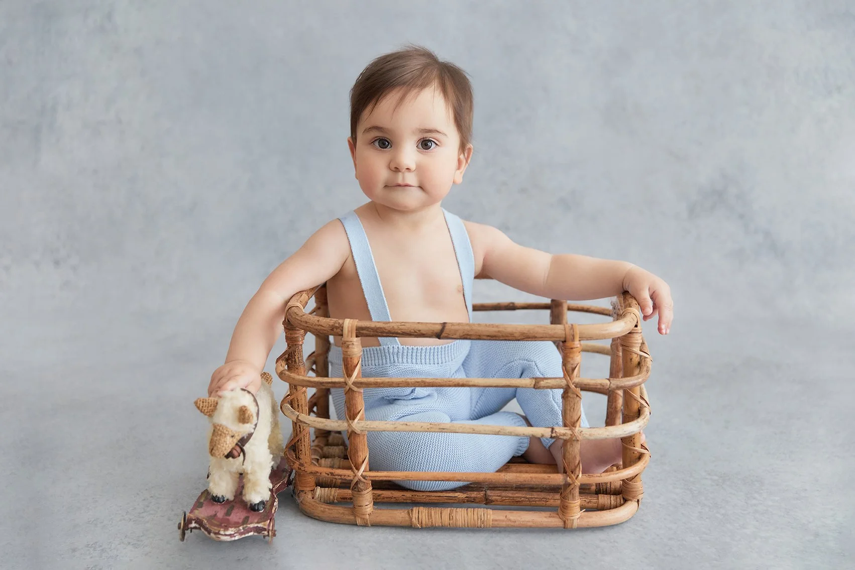 A baby with big eyes and brown hair sitting inside a wicker basket, holding a small stuffed animal with a party hat, against a plain light grey background.