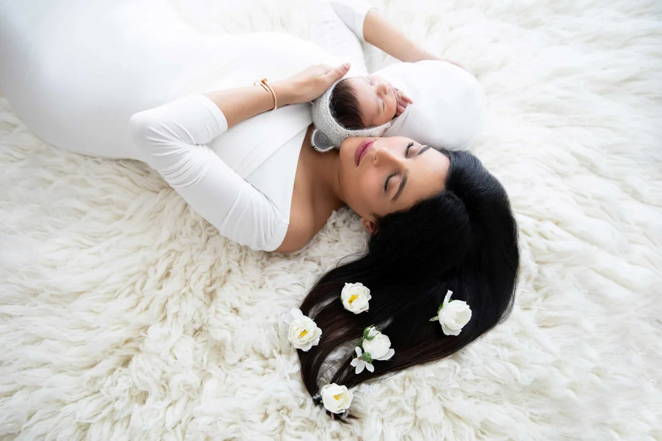 Mother lying on a fluffy white blanket with her eyes closed, holding her newborn baby wrapped in a blanket, with flowers in her long black hair.