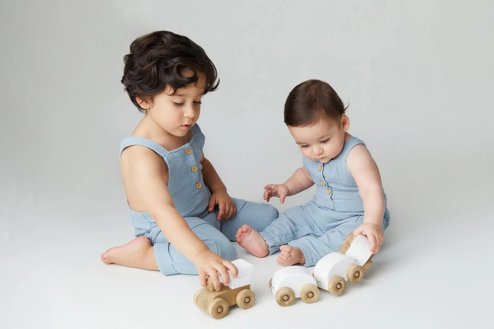 Two young children, a girl and a boy, sit on the floor playing with wooden toy trains. The children are wearing matching light blue outfits against a plain light background.