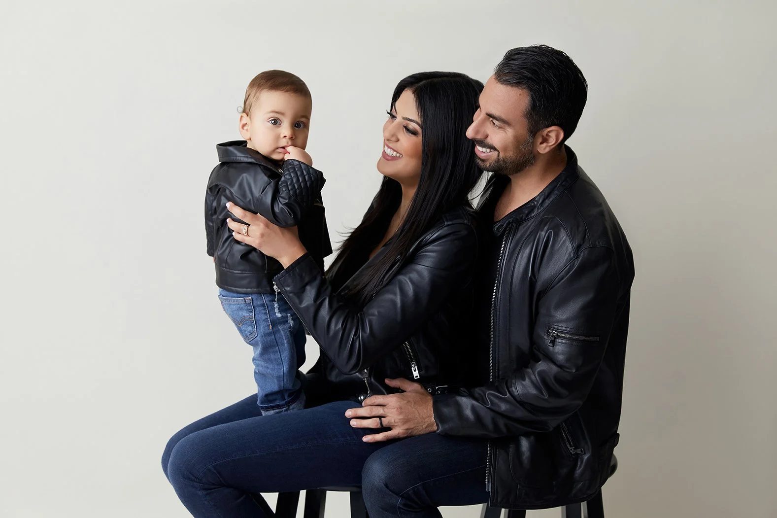 Family of three dressed in black leather jackets, the woman holding a young child, smiles, on a plain light background.