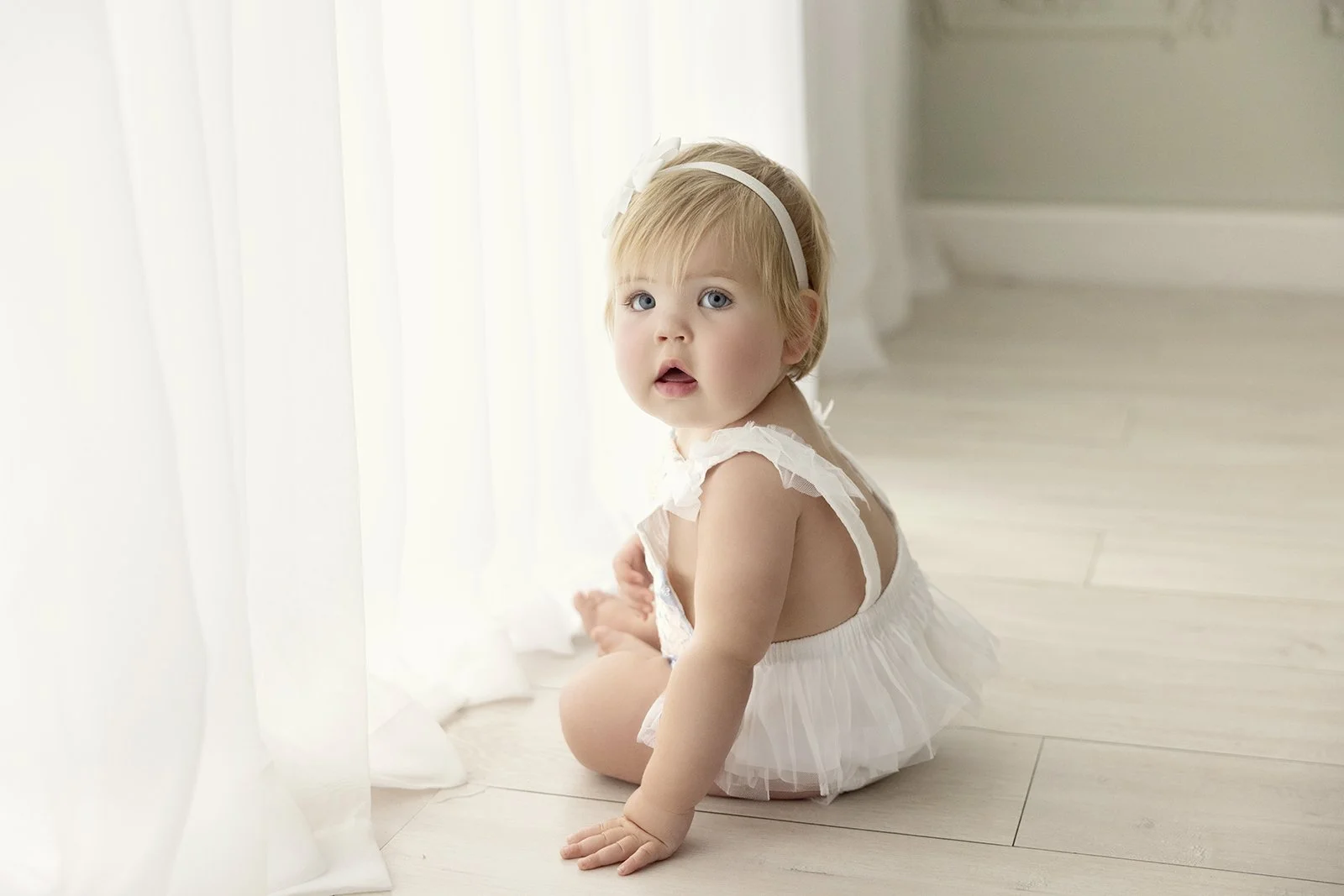 A young blonde girl in a white dress and headband sitting on wooden floor by a sheer curtain with a surprised expression.