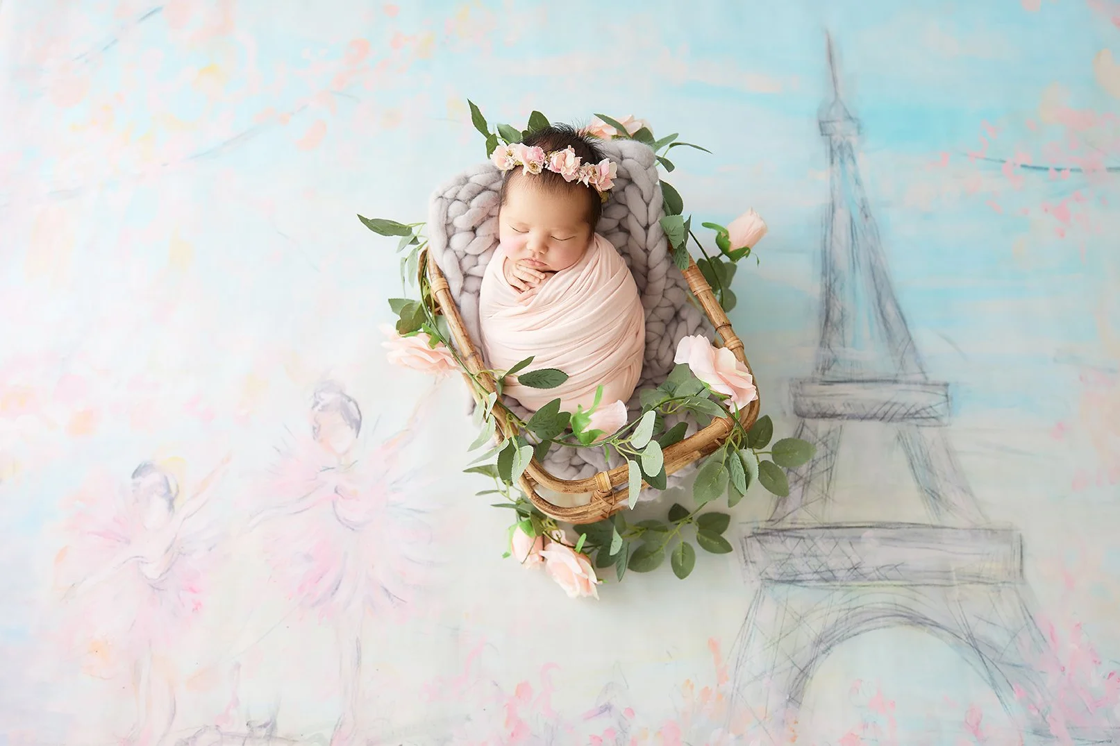 A sleeping baby swaddled in pink inside a wicker basket, adorned with pink roses and green leaves, against a pastel background with Paris and ballet dancer sketches.